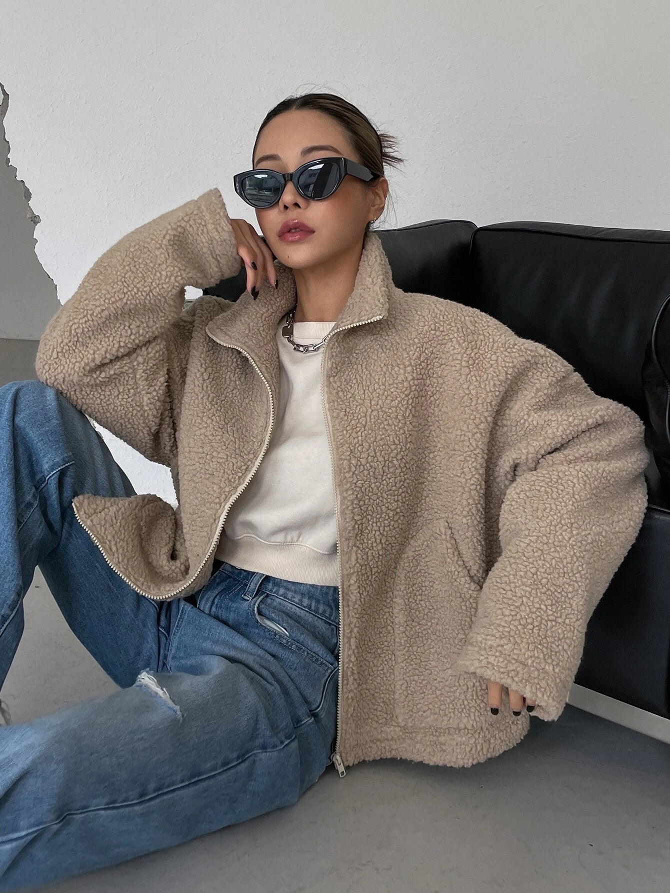 Frau in beiger Teddyjacke, lässig auf Sofa sitzend, trägt Sonnenbrille und Jeans. Modetrend, Herbstmode, Streetstyle, Damenbekleidung.