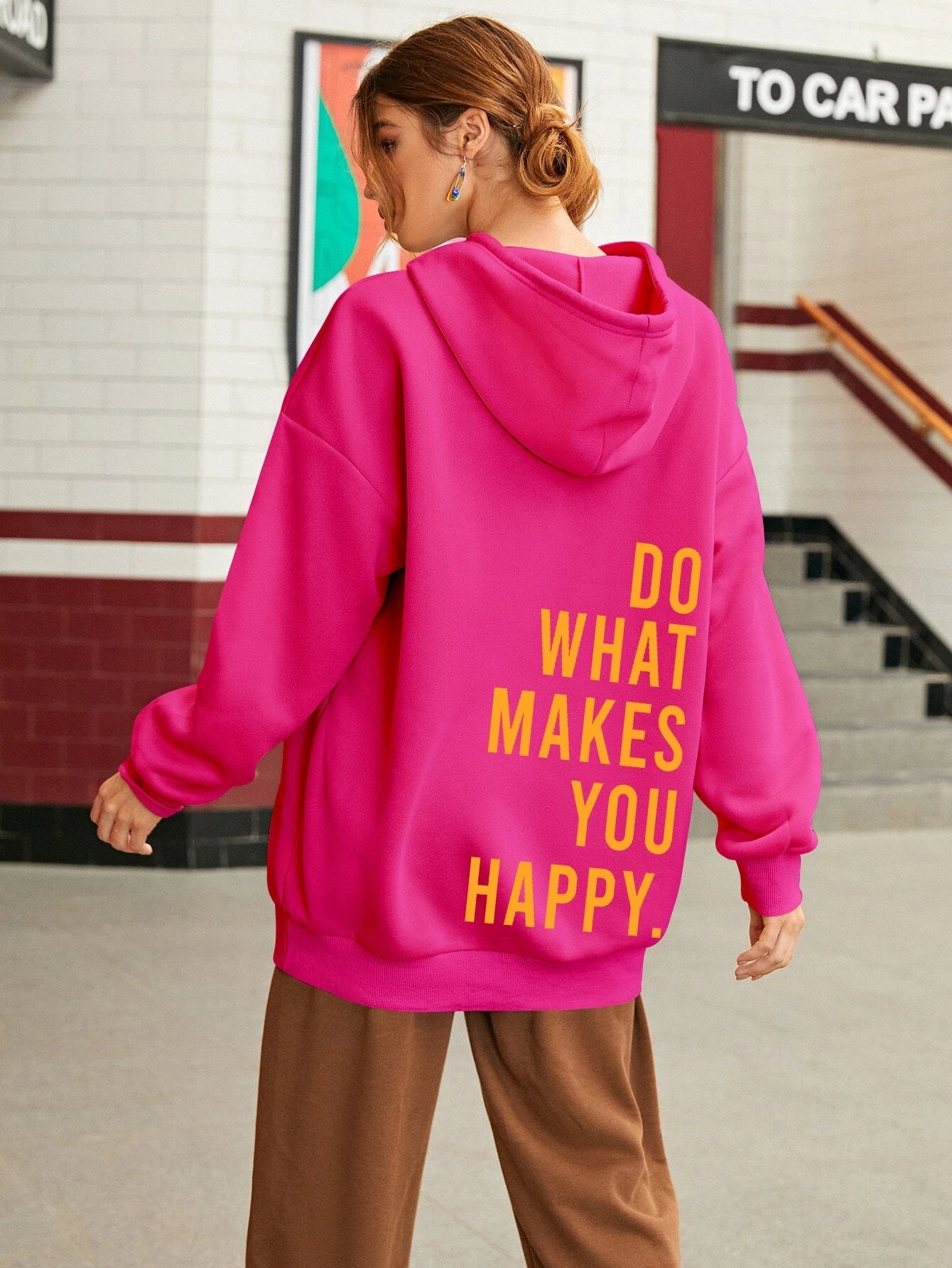 Frau in pinkem Hoodie mit gelbem Schriftzug "Do What Makes You Happy" in urbaner Umgebung. Modetrend, Streetwear, lässiger Stil.