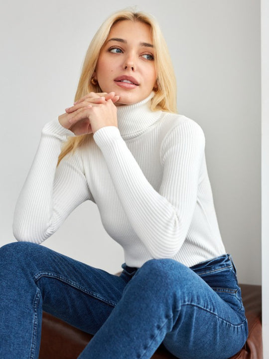 Blonde Frau in weißem Rollkragenpullover und blauer Jeans sitzt entspannt. Modetrend, Damenmode, stilvoll, lässig, Herbstoutfit.