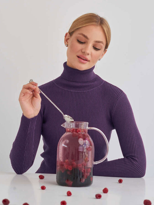 Frau in lila Rollkragenpullover rührt in Glaskaraffe mit Beeren und Getränk. Stilvoll, modern, gesund, Beerengetränk, Karaffe, Lifestyle.