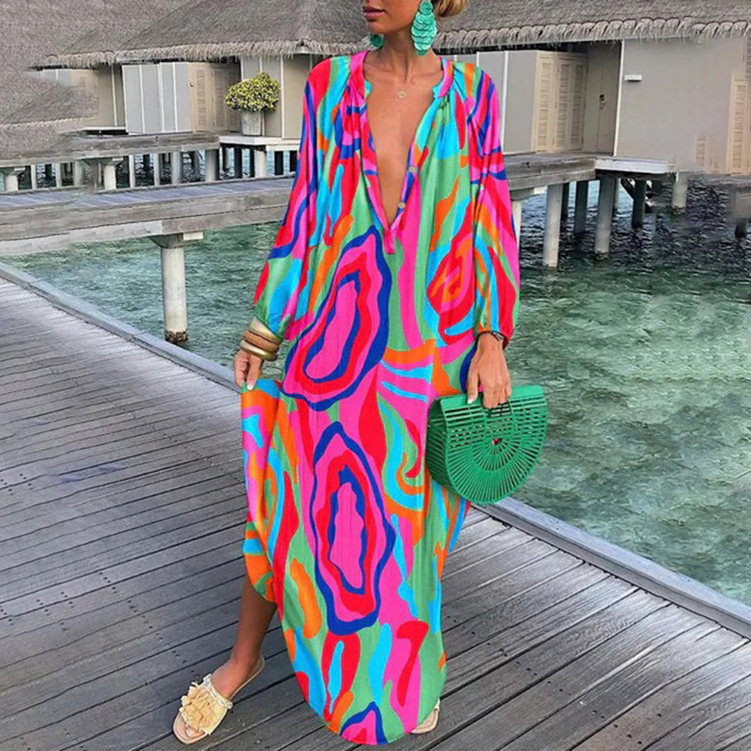 Frau in buntem, langem Kleid mit abstraktem Muster, steht auf Holzsteg am Wasser. Sommermode, Boho-Stil, leuchtende Farben, Strandkleidung.