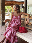 Frau in rotem Boho-Maxikleid mit Spaghettiträgern, sitzt auf Holzbank. Sommermode, Bohemian-Stil, elegantes Design, Outdoor-Hintergrund.