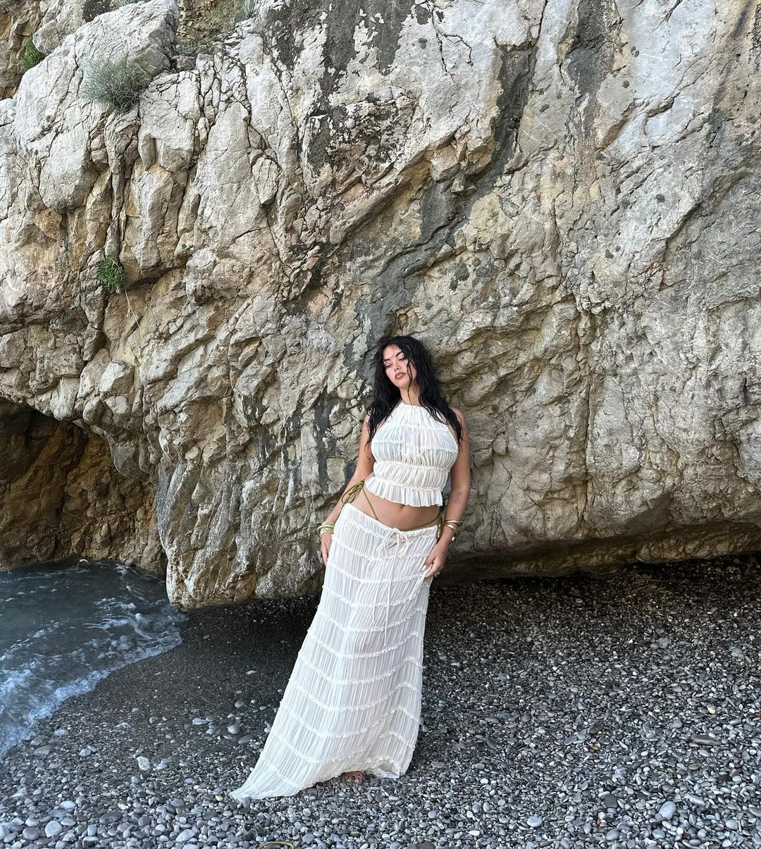 Frau in weißem Sommerkleid posiert vor einer felsigen Küste am Meer. Strandmode, Sommeroutfit, Naturkulisse, Urlaubsstimmung.