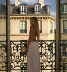 Frau in weißem Kleid auf Balkon mit kunstvollem Geländer, vor historischem Gebäude in Paris. Eleganz, Mode, Architektur, Stadtansicht.