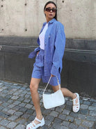 Frau in gestreiftem blauem Hemd und Shorts, weiße Sandalen, weiße Handtasche, Sonnenbrille. Modetrend, Sommeroutfit, Streetstyle.