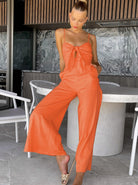 Frau in orangefarbenem Jumpsuit mit Schleifen-Detail, posiert stilvoll auf Terrasse. Sommermode, elegantes Design, trendige Damenbekleidung.