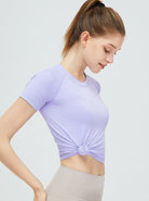 Frau in lila Sport-T-Shirt mit Knoten, ideal für Yoga und Fitness. Atmungsaktives Material, modisches Design, perfekte Passform für aktive Frauen.