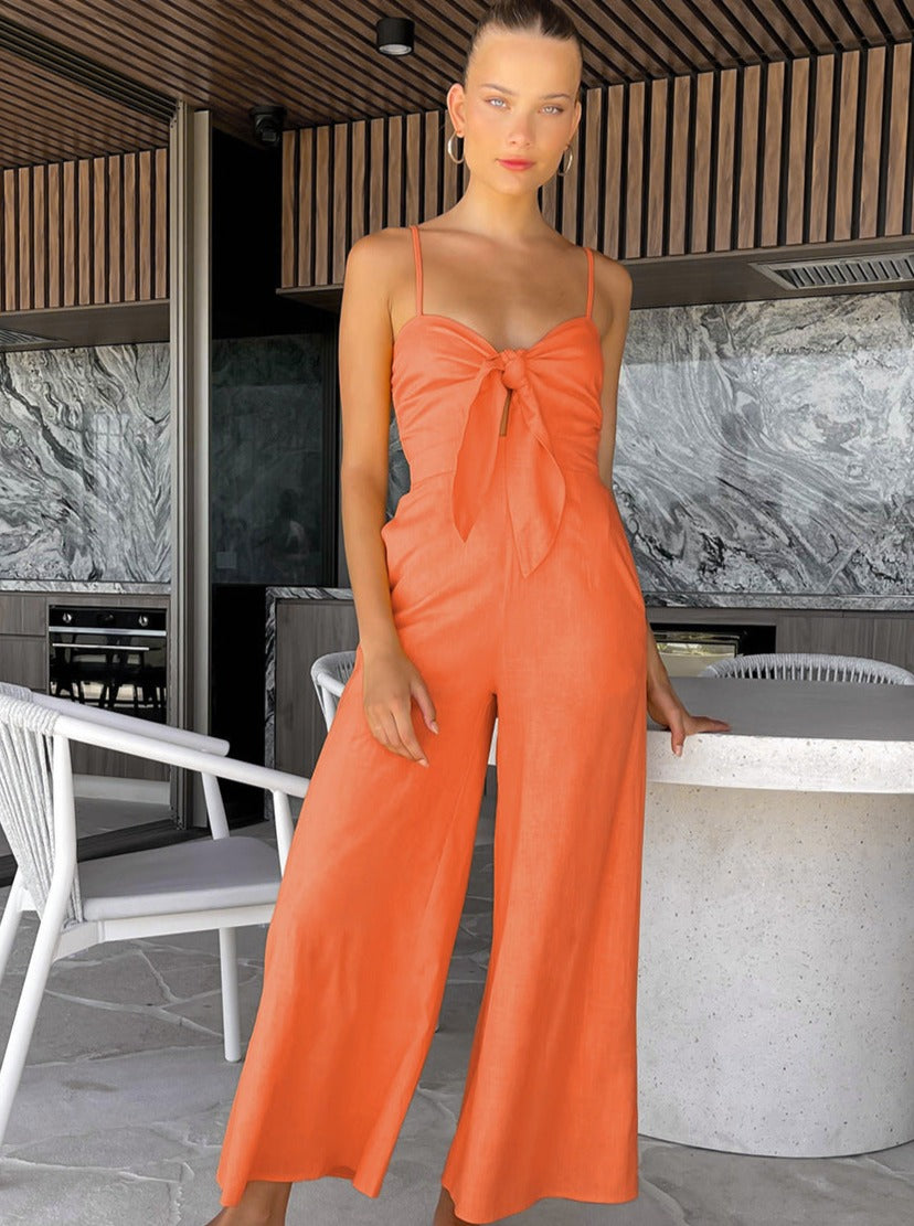 Frau in orangefarbenem Jumpsuit mit Schleifendetail, steht in modernem Innenraum. Sommermode, eleganter Look, trendige Damenbekleidung.