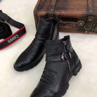 Schwarze Lederstiefel mit Reißverschluss und Schnalle, stilvoll und modern, neben einer Vintage-Kamera und einem Retro-Koffer auf weißem Hintergrund.