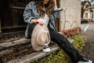 Frau in lässiger Jeansjacke und schwarzer Hose hält einen beigen Cowboyhut. Streetstyle, Mode, Herbstlook, lässige Kleidung, Outdoor-Fotografie.