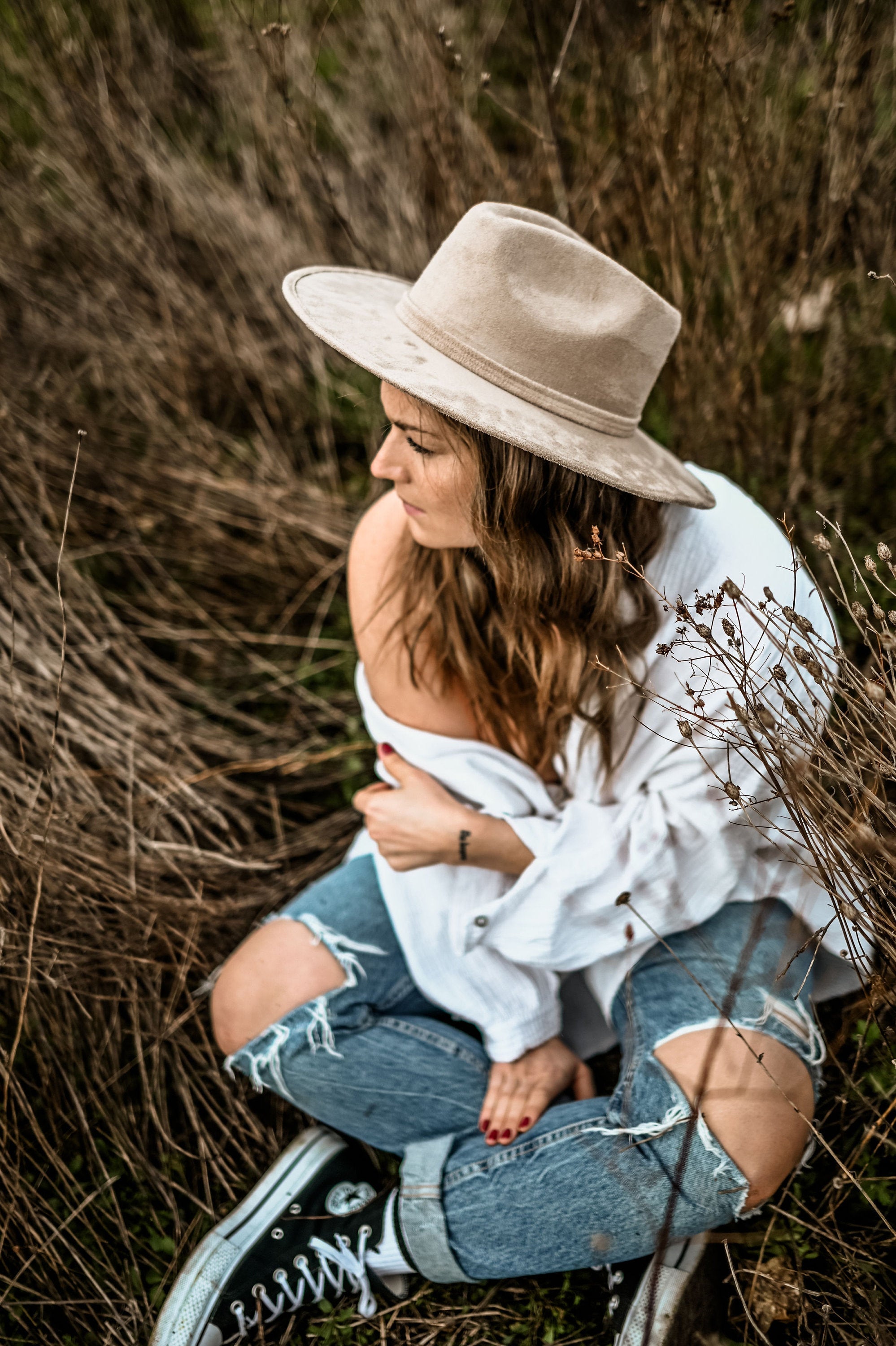 Frau in zerrissenen Jeans und weißem Hemd, sitzt im Gras, trägt einen beigen Hut. Mode, Outdoor, lässiger Stil, Herbstlook, Natur.