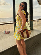 Frau in gelbem Sommerkleid mit Zitronenmuster, Sonnenbrille und Strohtasche posiert am Strandfenster. Perfektes Outfit für Sommermode am Meer.