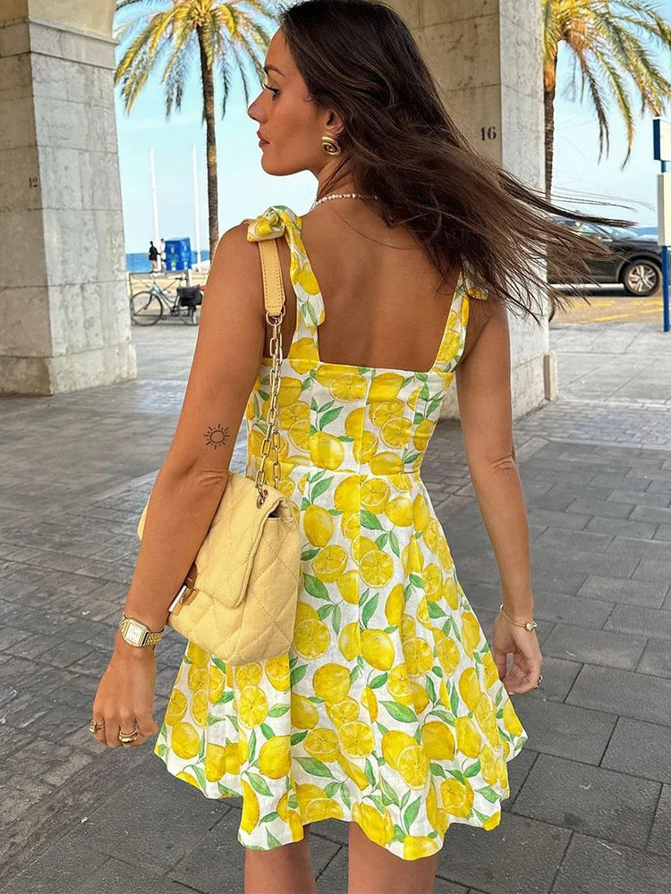 Frau im Sommerkleid mit Zitronenmuster, gelber Tasche, vor Palmenkulisse. Perfekt für Strandmode, Sommeroutfit, Urlaubslook.