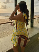 Frau in gelbem Sommerkleid mit Zitronenmuster blickt aufs Meer, trägt Strohtasche. Strandkulisse, Sonnenuntergang, Urlaubsstimmung.